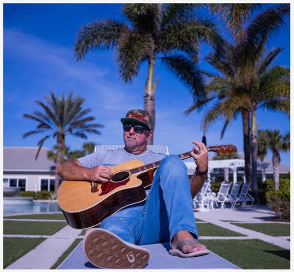 Man playing guitar outdoors by palm trees in casual attire.